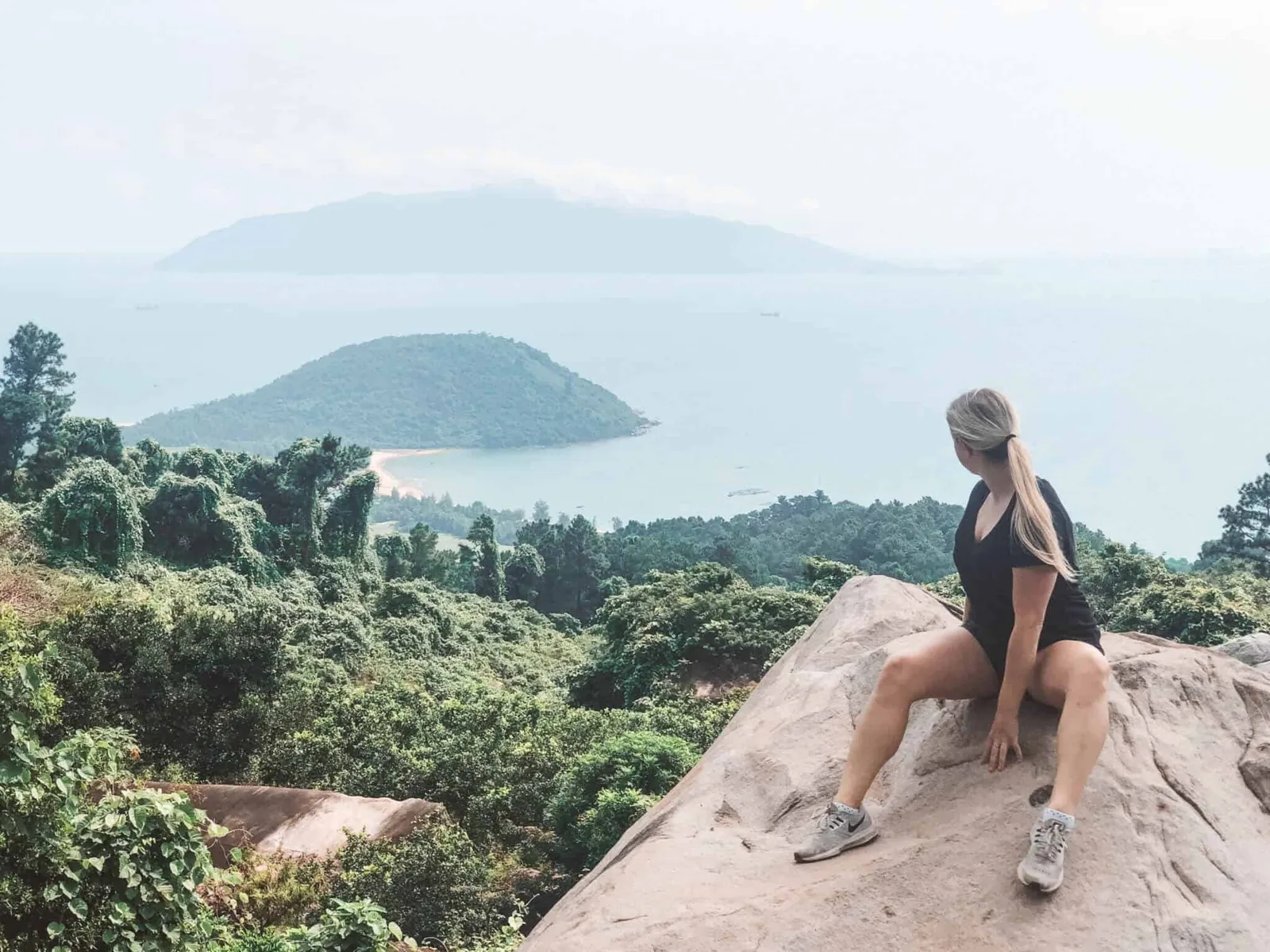 A woman sits on a rocky outcrop overlooking forested hills and islands along the coast near Hue.