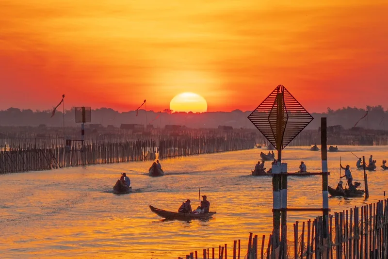 Small wooden boats glide across a river lagoon near Hue as the sun sets in a blazing orange sky.