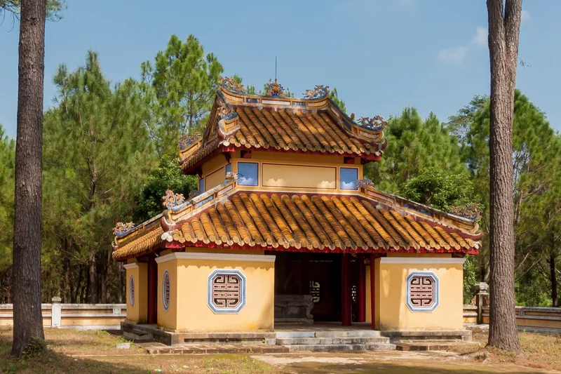 A yellow pavilion with ornate tiled roof stands amid pine trees at the Tomb of Emperor Gia Long in Hue.