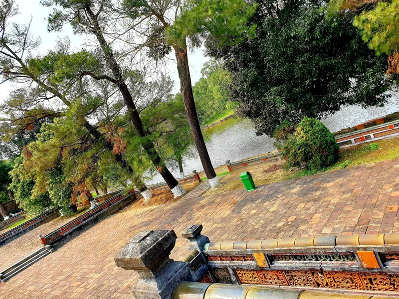 A tiled brick pathway lined with tall pine trees leads past a decorated stone balustrade beside a tranquil lake at Minh.